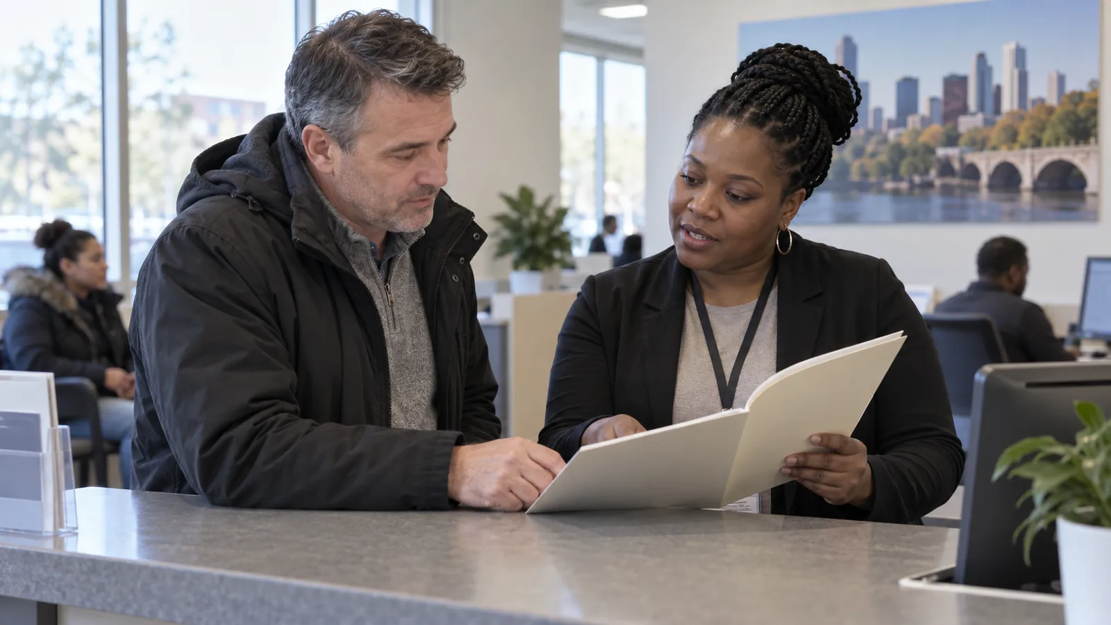 A visitor and a Black service navigator review a folder at a civic office counter.