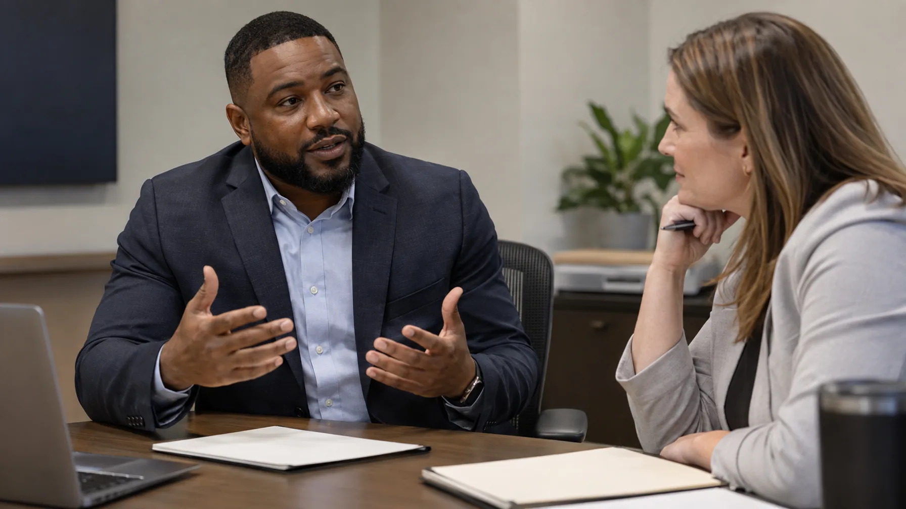 A professionally dressed Black man explains his perspective while a white woman colleague listens across a table.
