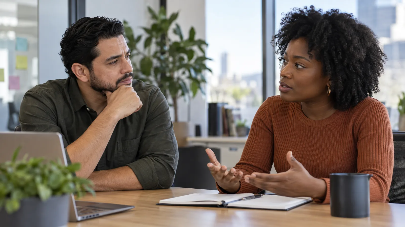 Two colleagues discuss a workplace interaction at a sunlit table.